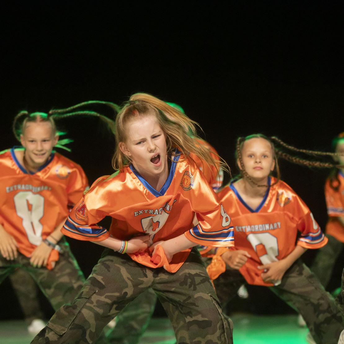 M&auml;dchen in orangenen Sporttrikots tanzen leidenschaftlich auf der B&uuml;hne, umgeben von weiteren T&auml;nzerinnen in Camouflage-Hosen.