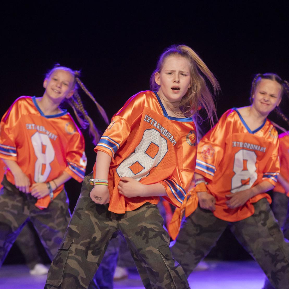 Tanzende Gruppe von M&auml;dchen in orangefarbener Sportbekleidung mit Camouflage-Hosen auf einer B&uuml;hne.