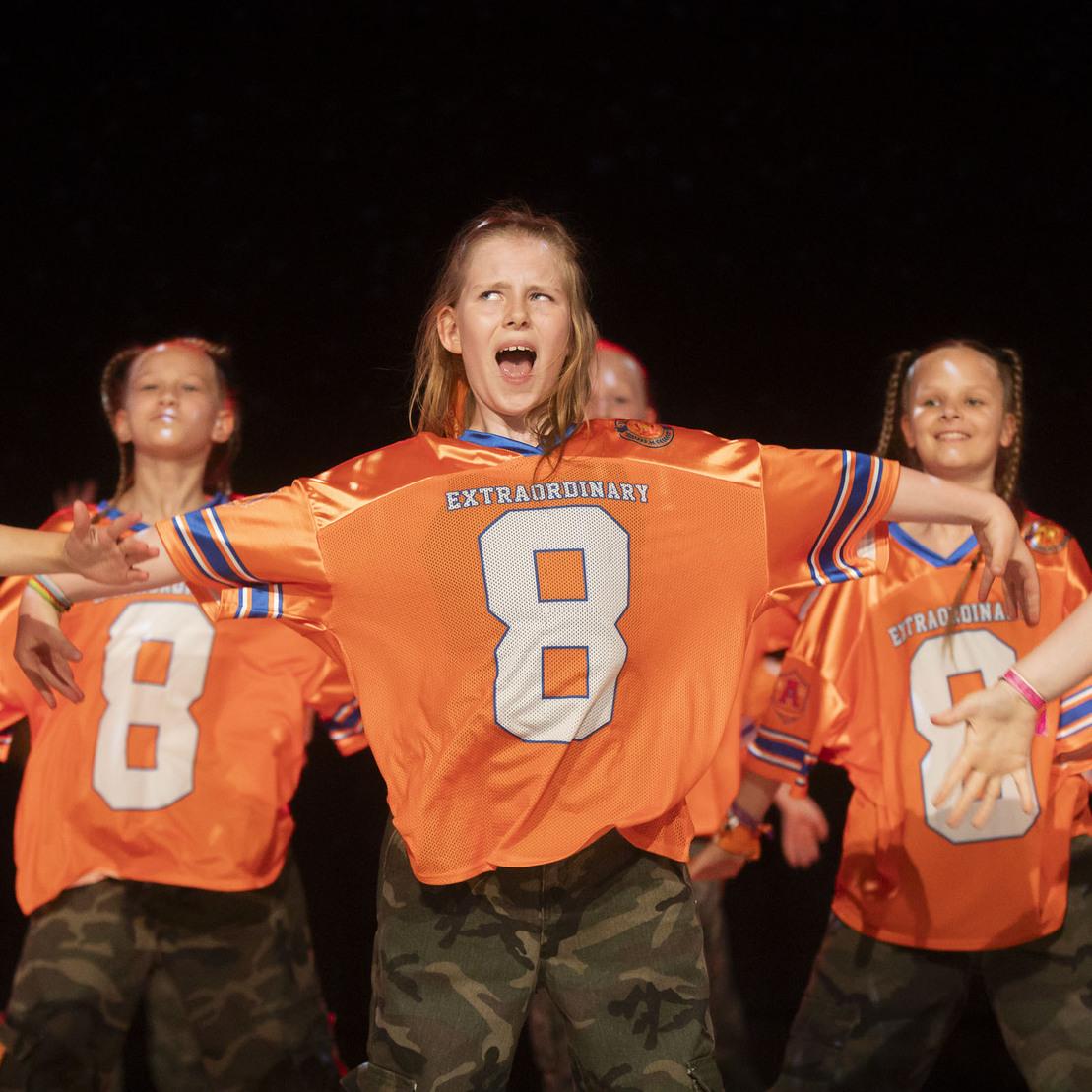 Tanzende Kinder in orangefarbenen Trikots mit der Nummer 8 und Camouflage-Hosen pr&auml;sentieren eine lebhafte Choreografie.