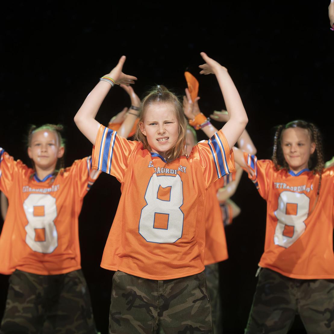 M&auml;dchen in orangefarbenen Trikots mit der Nummer 8 tanzen energetisch auf einer B&uuml;hne.