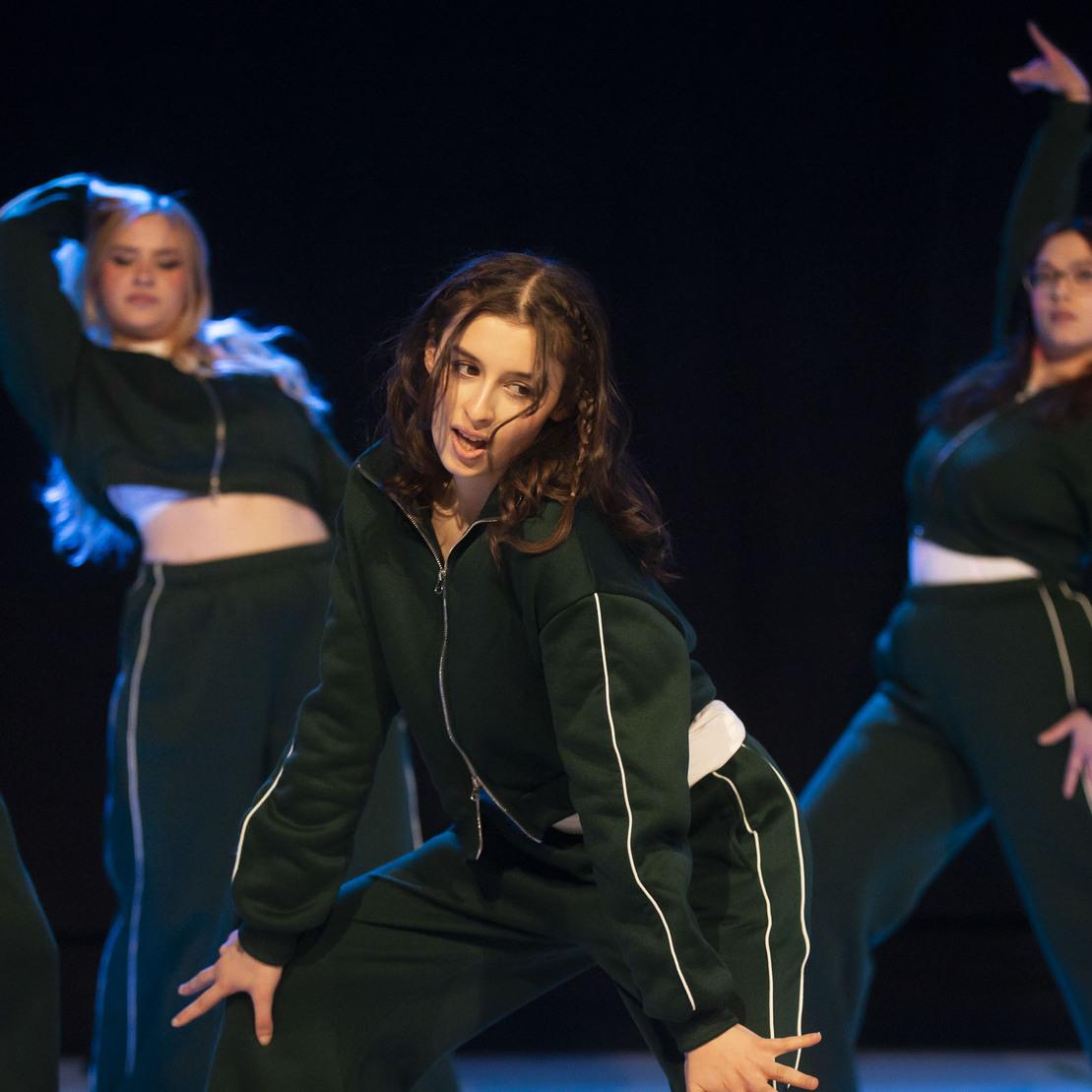 Vier T&auml;nzerinnen in gr&uuml;nen Sportoutfits performen auf einer B&uuml;hne, eine im Vordergrund hebt ihre H&auml;nde kreativ.