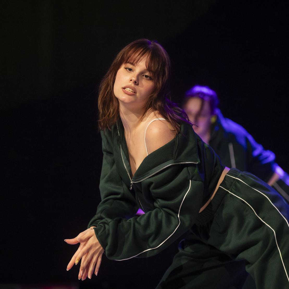T&auml;nzerin in gr&uuml;ner Sportbekleidung bewegt sich ausdrucksstark auf einer B&uuml;hne mit bunten Lichtern im Hintergrund.