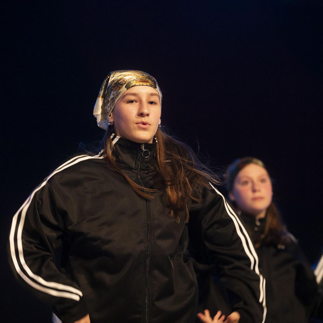 T&auml;nzerin in schwarzer Trainingsjacke mit Kopftuch, w&auml;hrend einer Performance auf der B&uuml;hne, von weiteren T&auml;nzerinnen umgeben.