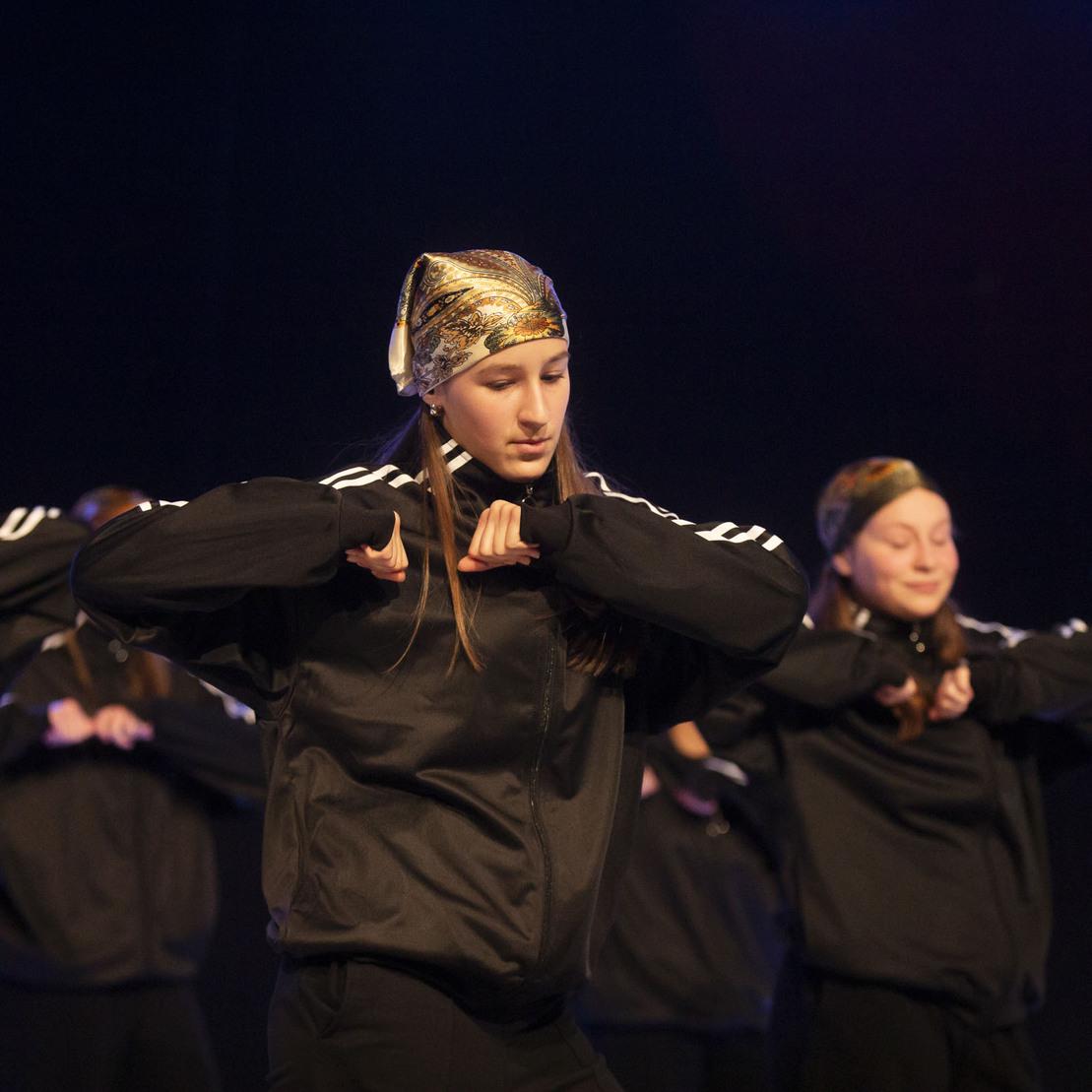 T&auml;nzerinnen in schwarzen Trainingsanz&uuml;gen und Kopfbedeckungen pr&auml;sentieren synchronisierte Bewegungen auf einer B&uuml;hne.
