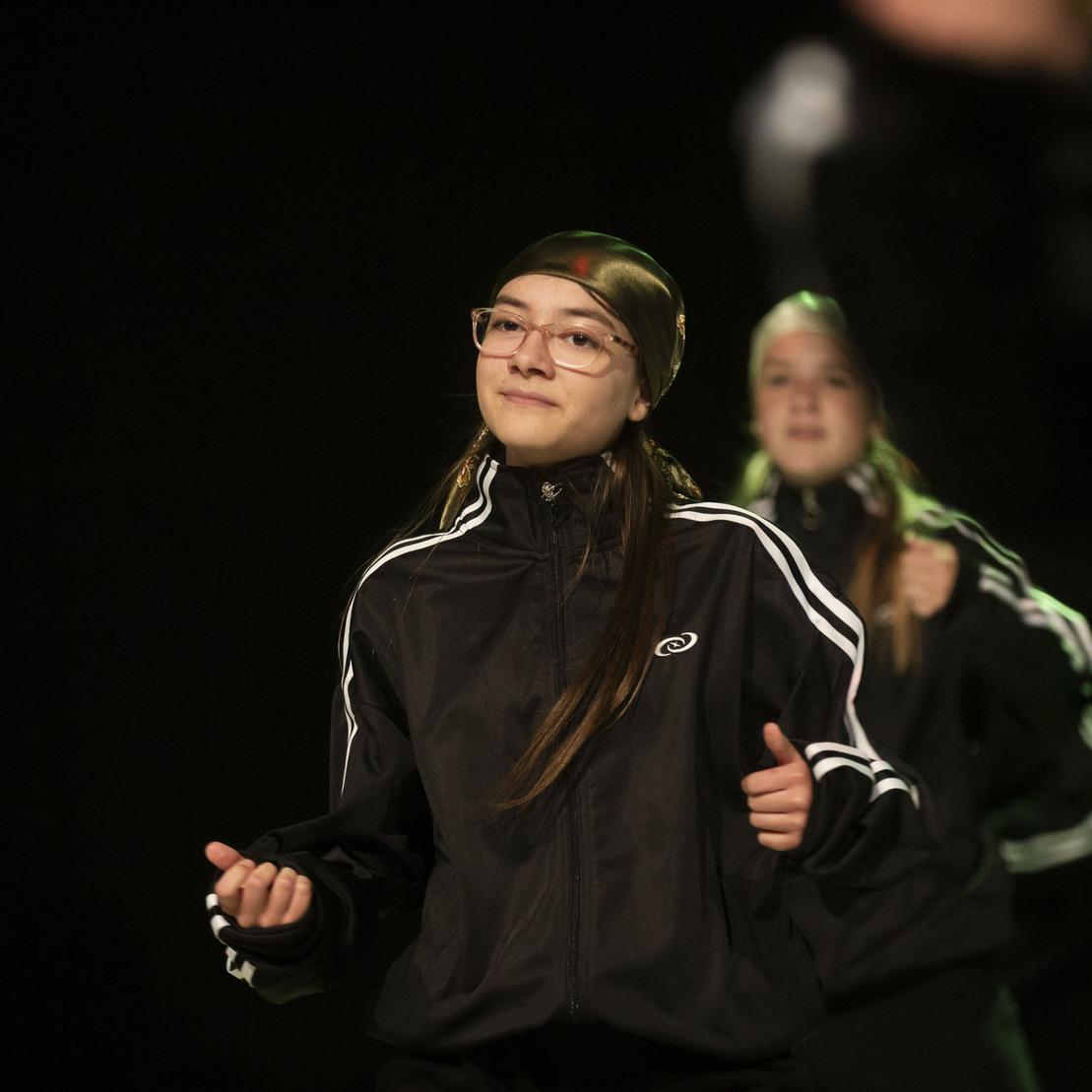 T&auml;nzerin in schwarzer Sportjacke mit Brille, die auf einer B&uuml;hne eine choreografierte Bewegung ausf&uuml;hrt.
