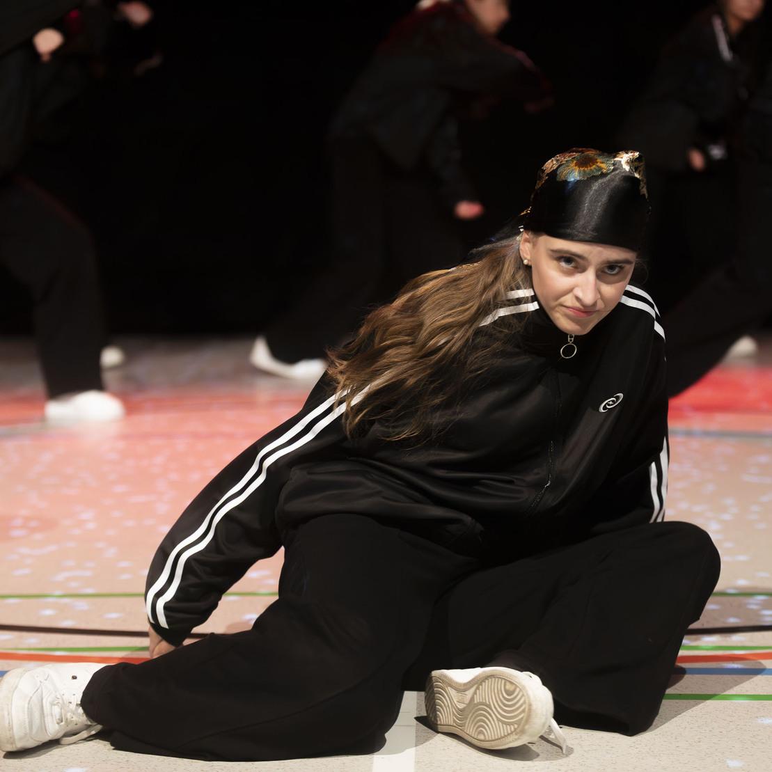 T&auml;nzerin in schwarzem Outfit sitzt auf dem Boden, w&auml;hrend andere T&auml;nzer im Hintergrund in Bewegung sind.