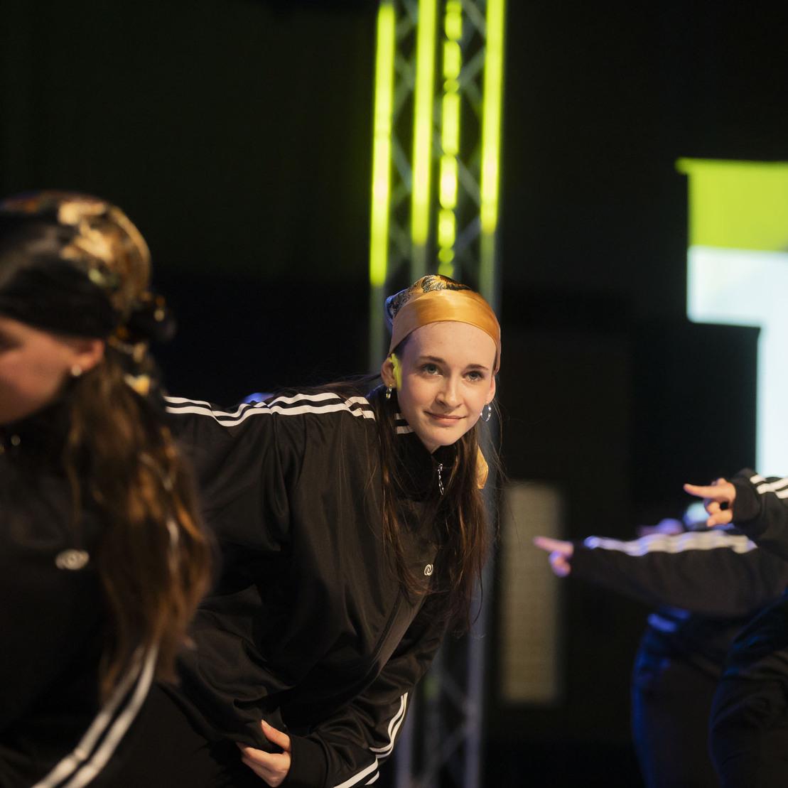 Tanzgruppe in schwarzen Outfits f&uuml;hrt eine Choreografie auf einer B&uuml;hne mit bunten Lichtern vor.