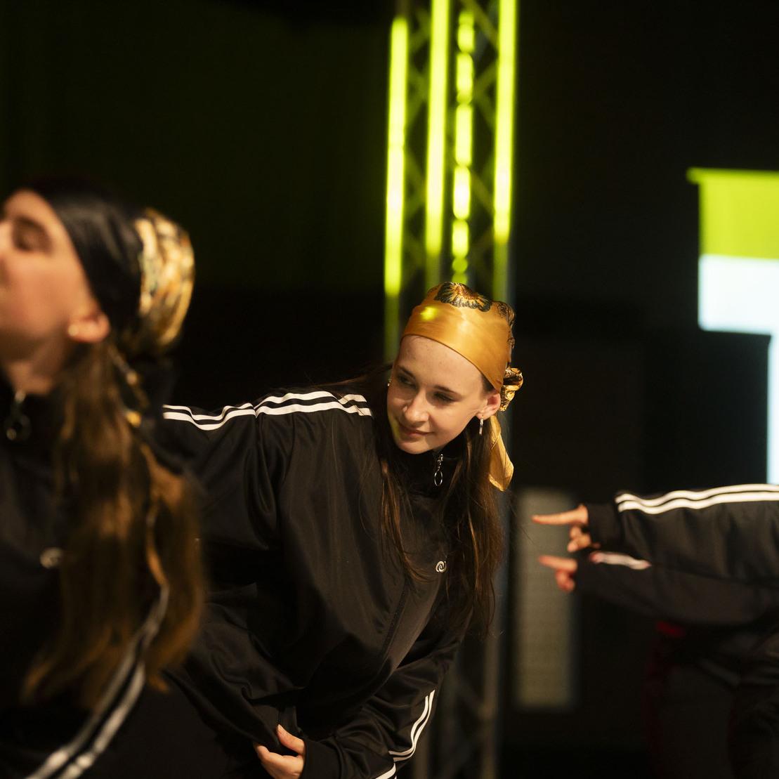 Gruppentanz von Jugendlichen in sportlicher Kleidung auf einer B&uuml;hne, mit Fokus auf zwei T&auml;nzerinnen.