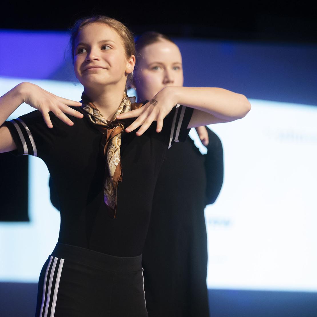 Zwei T&auml;nzerinnen pr&auml;sentieren eine Choreografie auf der B&uuml;hne, eine mit einem Halstuch, w&auml;hrend ein projizierter Hintergrund sichtbar ist.