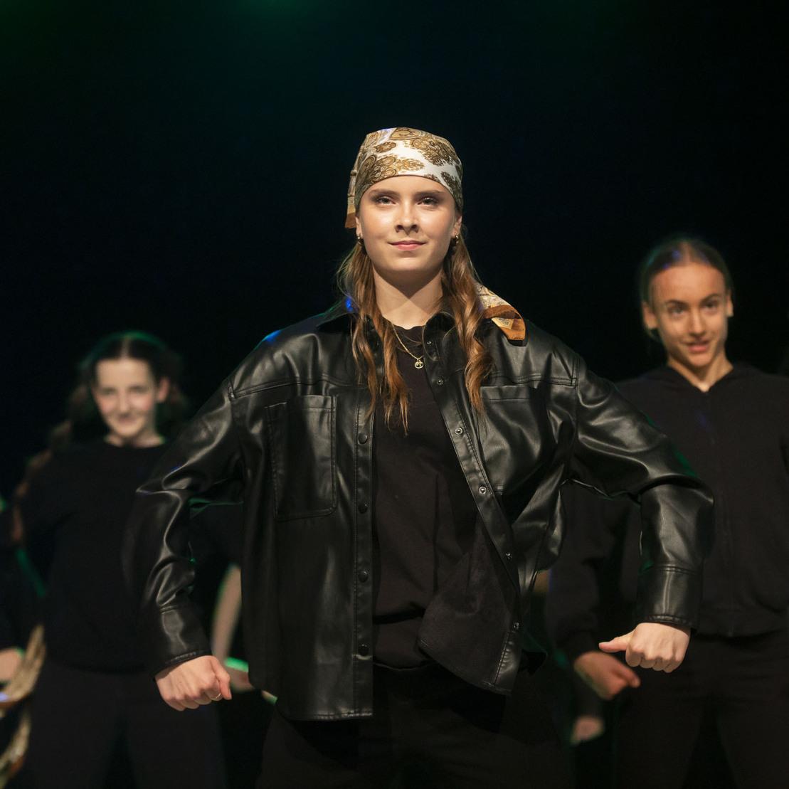 T&auml;nzerin in Lederjacke mit Bandana steht im Vordergrund einer Gruppe, die auf einer B&uuml;hne performt.