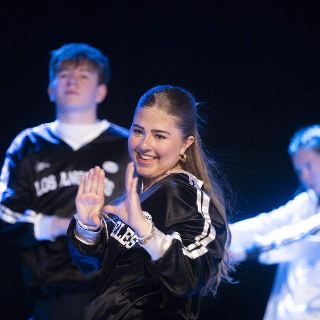 Drei T&auml;nzer in Trainingsanz&uuml;gen performen auf einer B&uuml;hne mit blauem Licht. Die vordere T&auml;nzerin l&auml;chelt und gestikuliert.