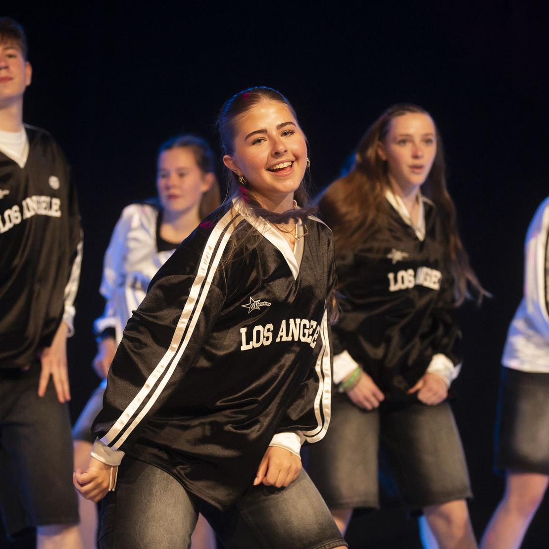 Tanzgruppe in sportlichen Outfits f&uuml;hrt auf einer B&uuml;hne eine Choreografie mit L&auml;cheln und Energie auf.