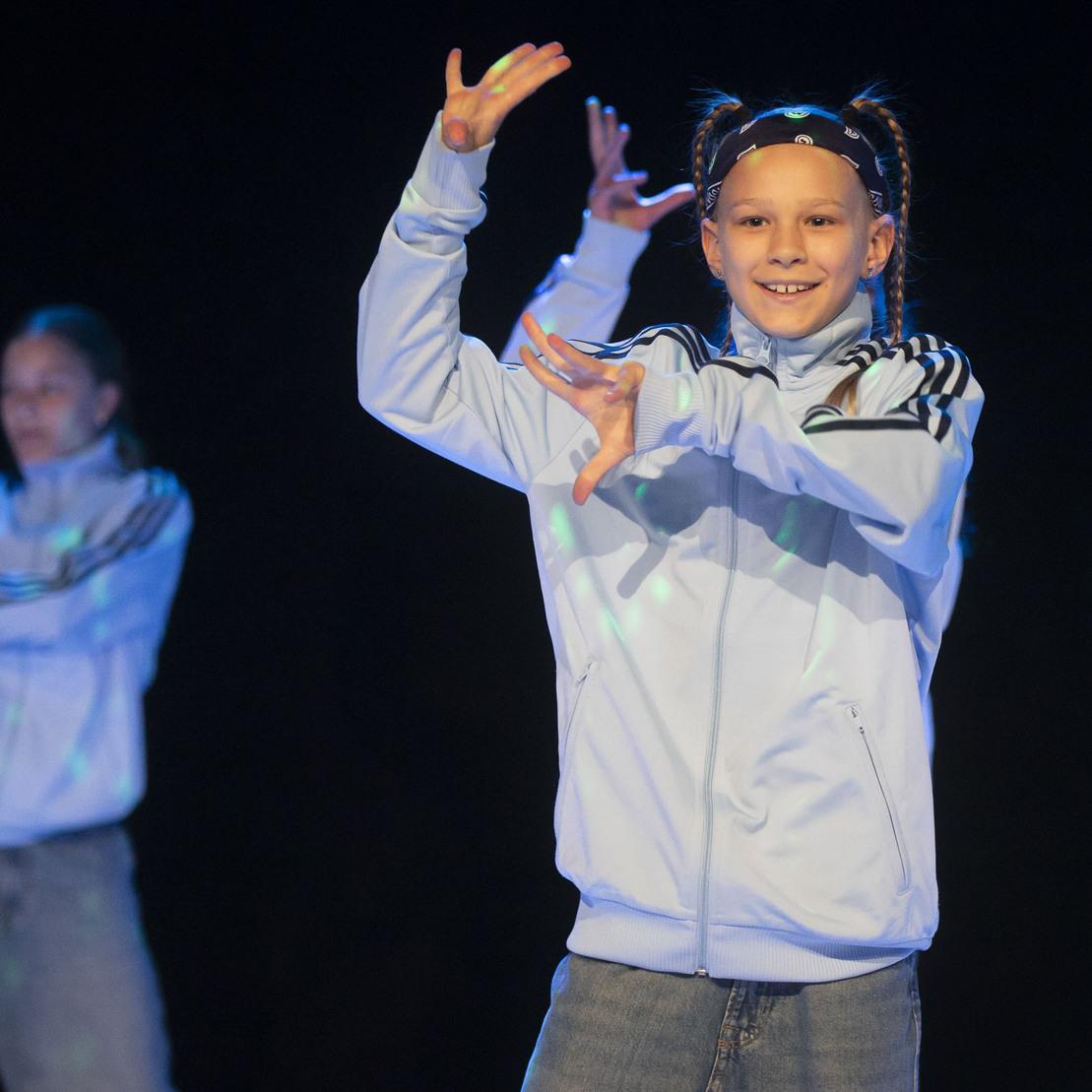 L&auml;chelndes Kind in blauer Sportjacke tanzt mit einem anderen T&auml;nzer im Hintergrund auf einer B&uuml;hne mit bunten Lichtern.
