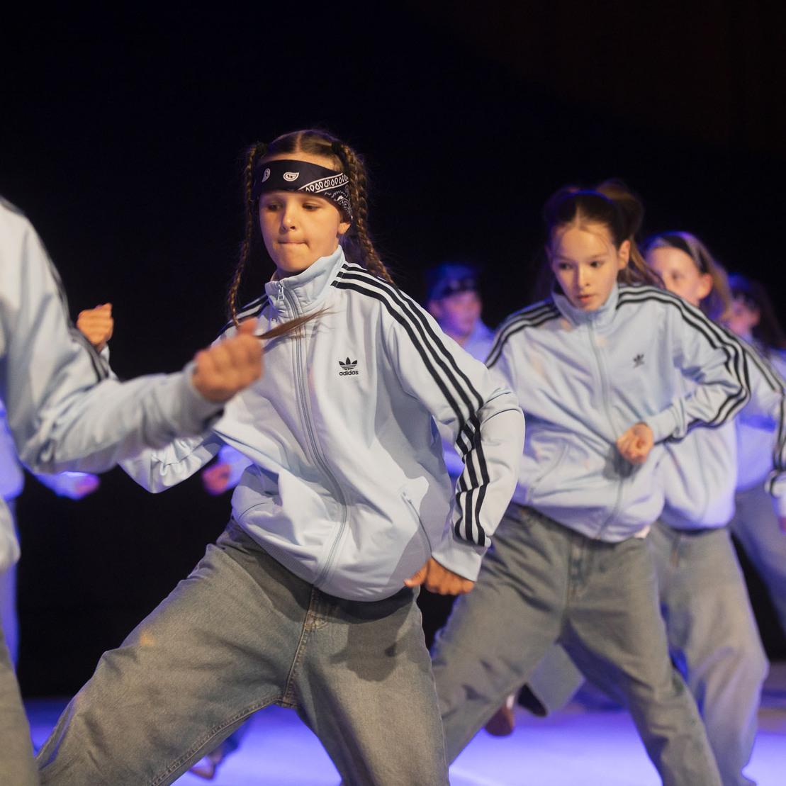 Tanzgruppe in blauen Adidas-Jacken und Jeans f&uuml;hrt synchronisierte Bewegungen auf einer B&uuml;hne aus.
