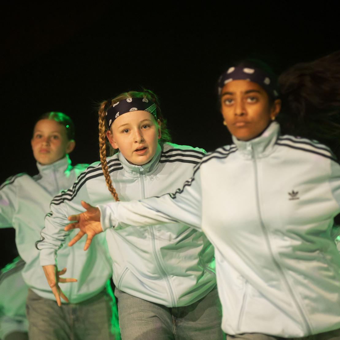 Tanzgruppe in blauen Jacken f&uuml;hrt eine Choreografie auf einer B&uuml;hne mit gr&uuml;nem Licht aus.