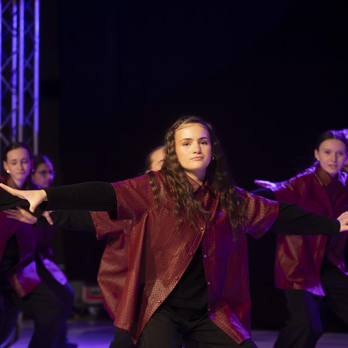 T&auml;nzerinnen in roten Oberteilen f&uuml;hren eine Choreografie auf einer B&uuml;hne mit Scheinwerferlicht auf.