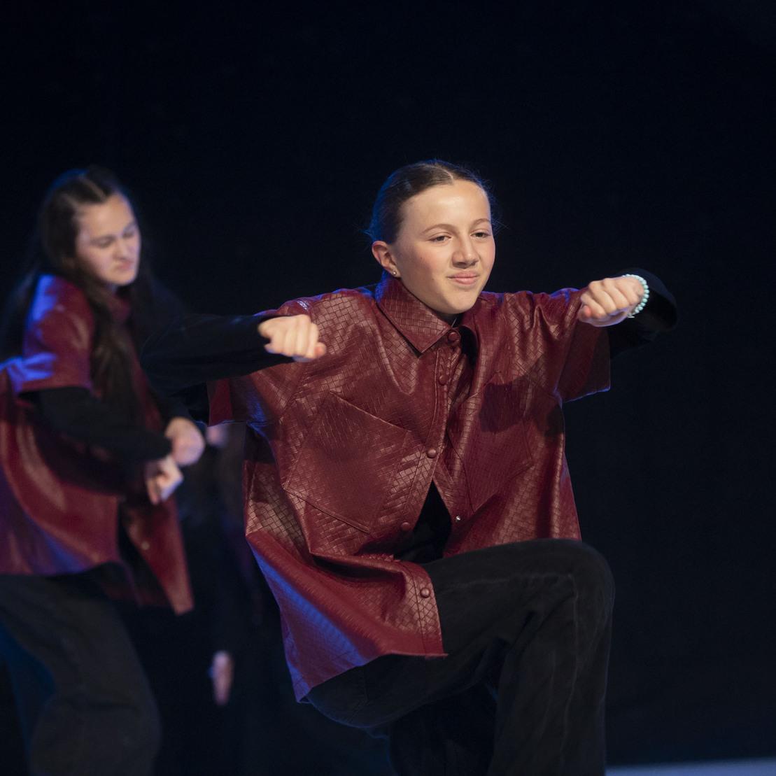 T&auml;nzerinnen in roter Kleidung zeigen eine choreografierte Bewegung auf einer dunklen B&uuml;hne.