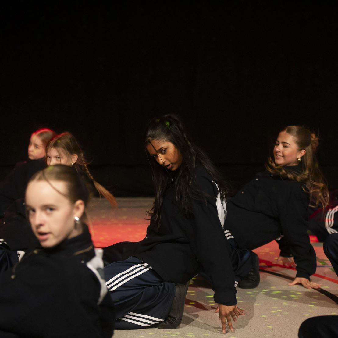 Gruppe von T&auml;nzerinnen in sportlichen Outfits f&uuml;hrt eine choreografierte Tanzroutine auf einer beleuchteten B&uuml;hne aus.