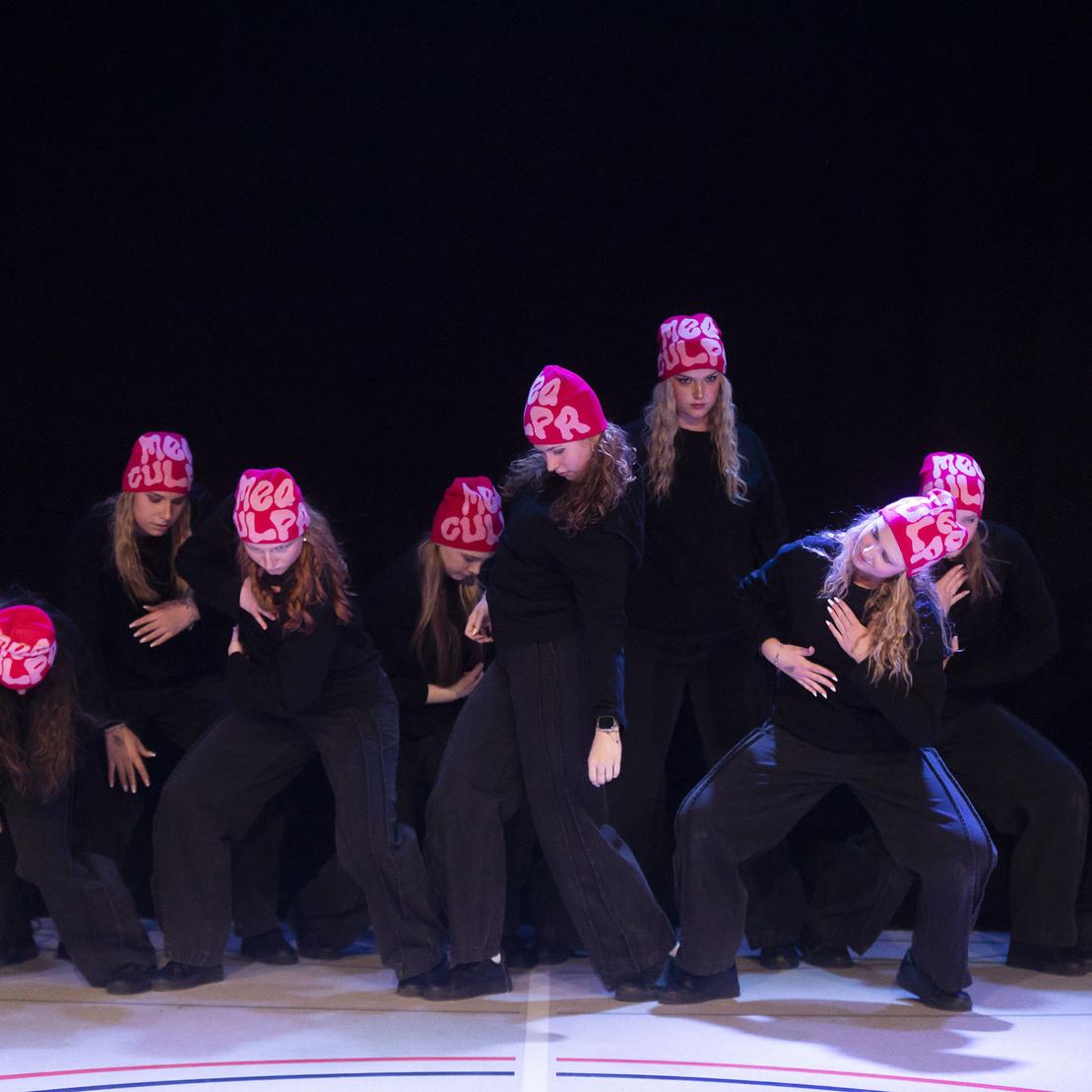 Tanzgruppe in schwarzer Kleidung mit pinken M&uuml;tzen posiert dynamisch auf einer B&uuml;hne.