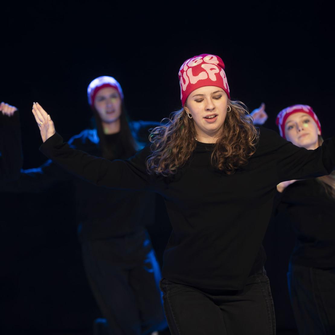 T&auml;nzerinnen in schwarzen Outfits und bunten M&uuml;tzen f&uuml;hren eine choreografierte Tanzroutine auf einer dunklen B&uuml;hne vor.