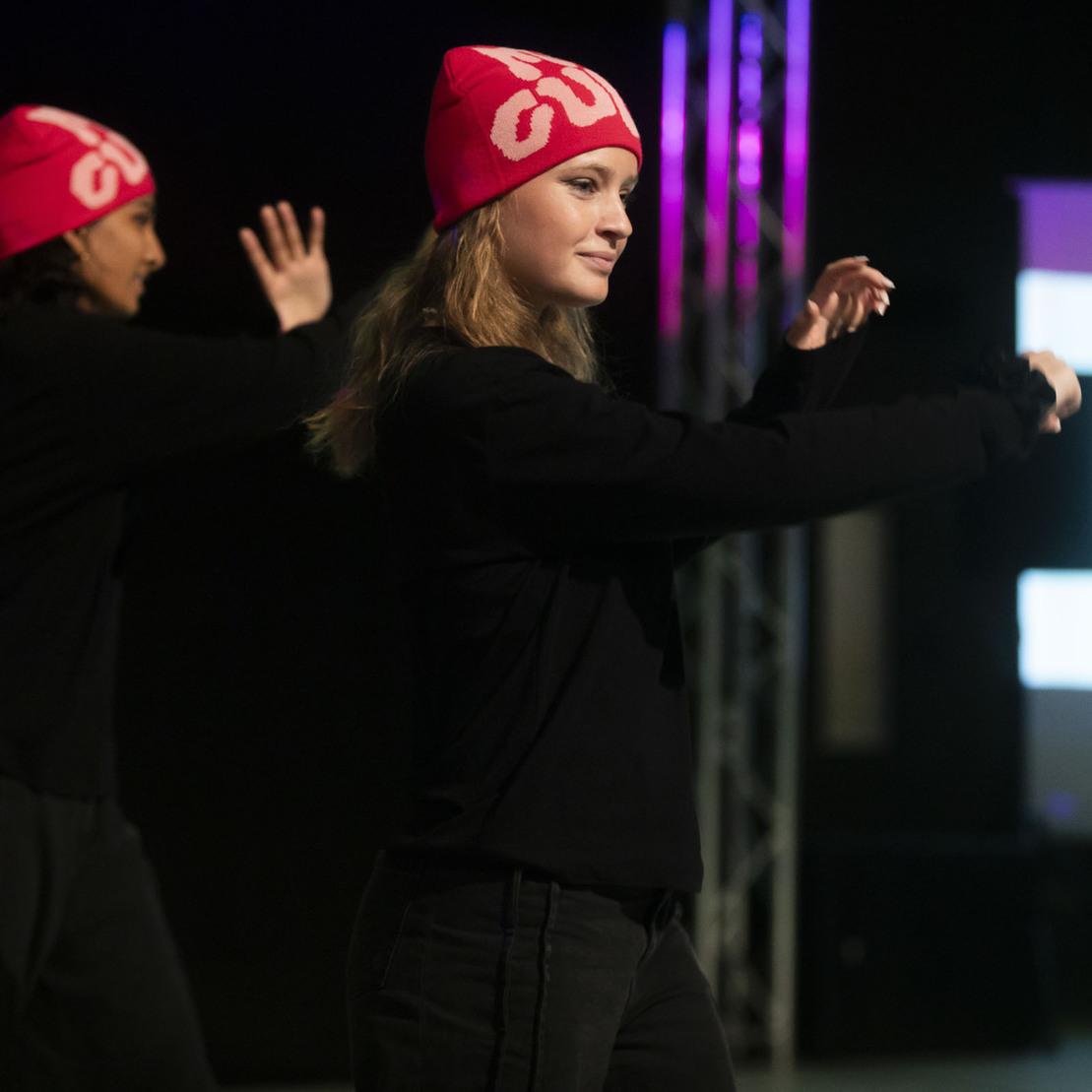 Zwei T&auml;nzerinnen in schwarzen Oberteilen und pinken M&uuml;tzen f&uuml;hren synchron eine Choreografie auf einer B&uuml;hne vor.
