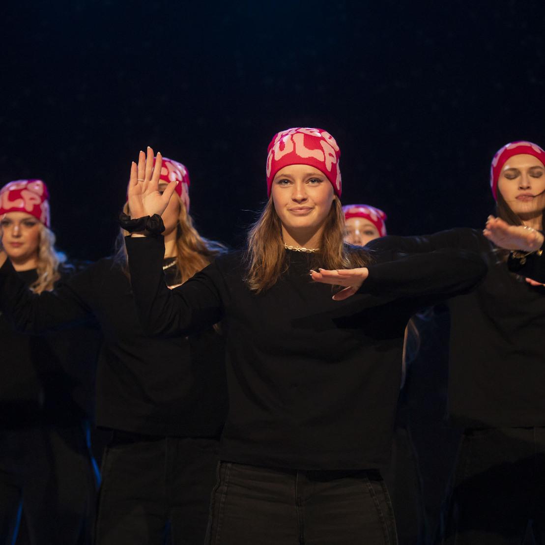 Tanzgruppe mit mehreren Frauen in schwarzen Outfits und pinken M&uuml;tzen, die synchron Bewegungen ausf&uuml;hren.