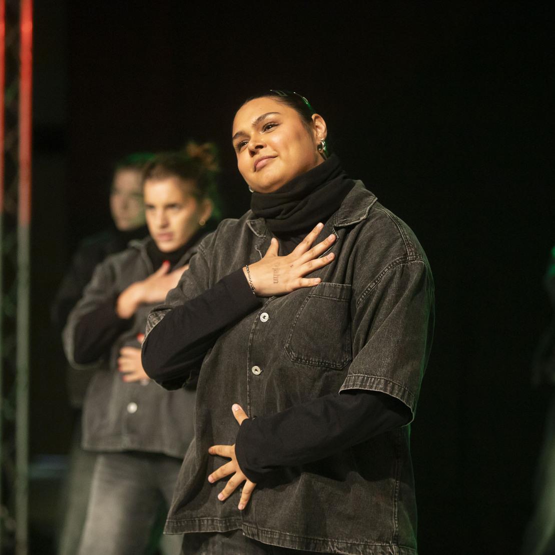 Tanzende Gruppe auf der B&uuml;hne, eine T&auml;nzerin mit erhobener Hand in einer Denimjacke, im Vordergrund sichtbar.