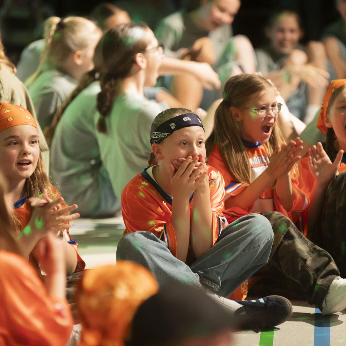 Gruppe von Kindern in orangefarbenen Shirts und Bandanas, die begeistert bei einer Veranstaltung sitzen und jubeln.