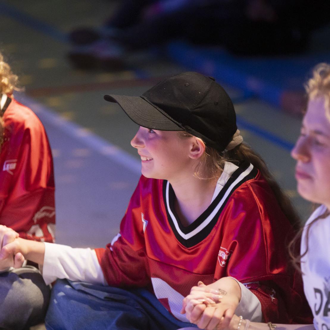 Drei Jugendliche in Sporttrikots sitzen im Kreis und halten sich an den H&auml;nden, l&auml;chelnd auf einem Boden belassenen Raum.