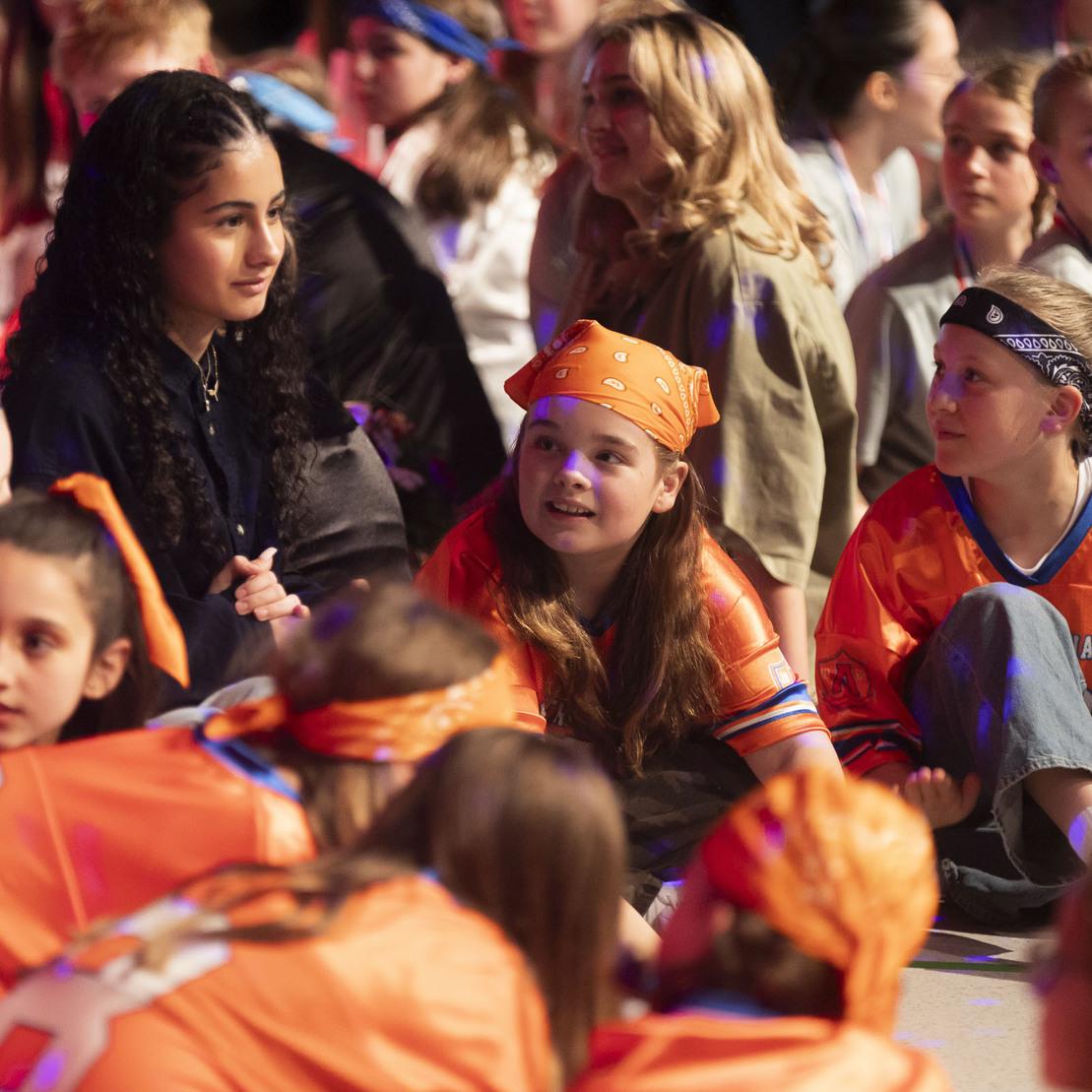 Gruppenszene mit Kindern in orangefarbener Kleidung und Gesichtst&uuml;chern, die miteinander sprechen und lachen.