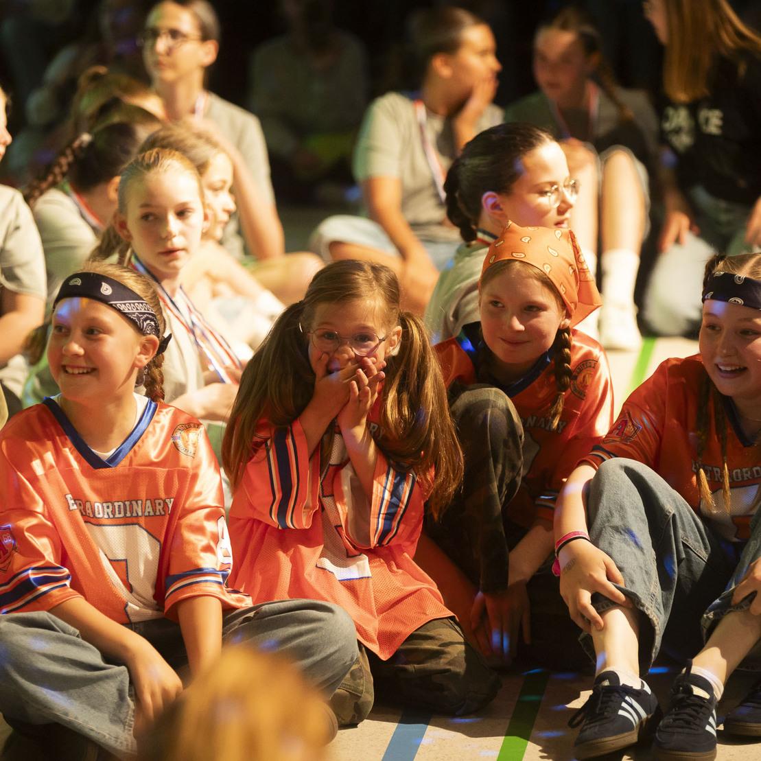 Gruppe von Kindern in sportlicher Kleidung sitzt l&auml;chelnd und aufmerksam auf dem Boden w&auml;hrend einer Veranstaltung.