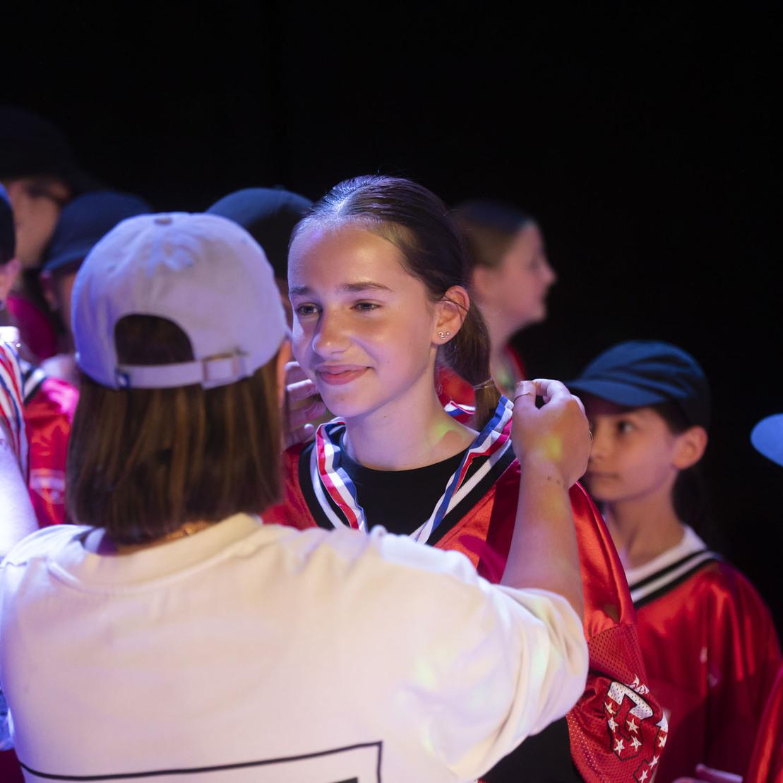 Ein M&auml;dchen erh&auml;lt eine Medaille von einer Person, w&auml;hrend weitere Kinder in roten Trikots im Hintergrund warten.