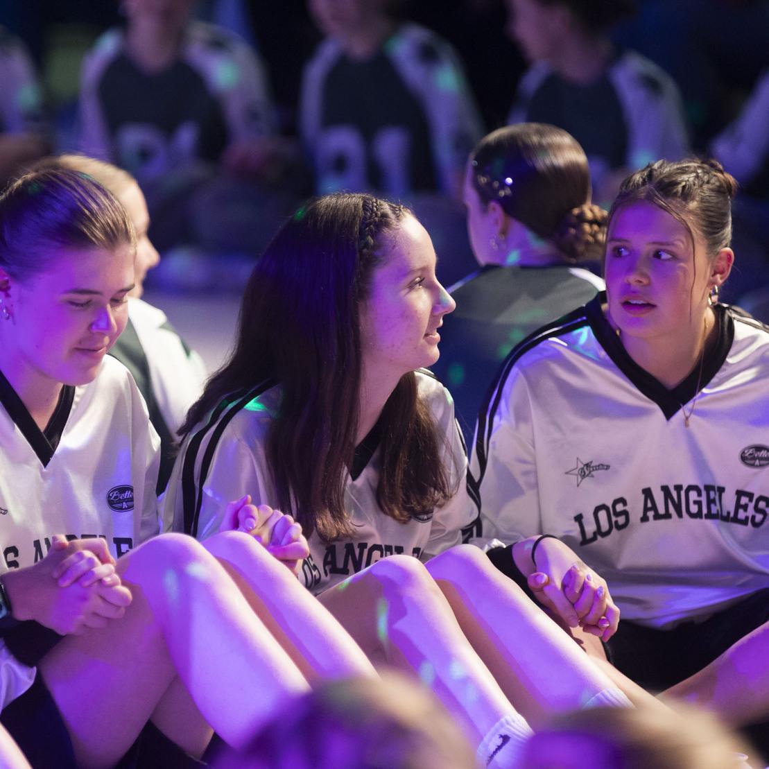 Drei M&auml;dchen in wei&szlig;en Sporttrikots mit &bdquo;LOS ANGELES&ldquo;-Aufdruck sitzen auf dem Boden und unterhalten sich.