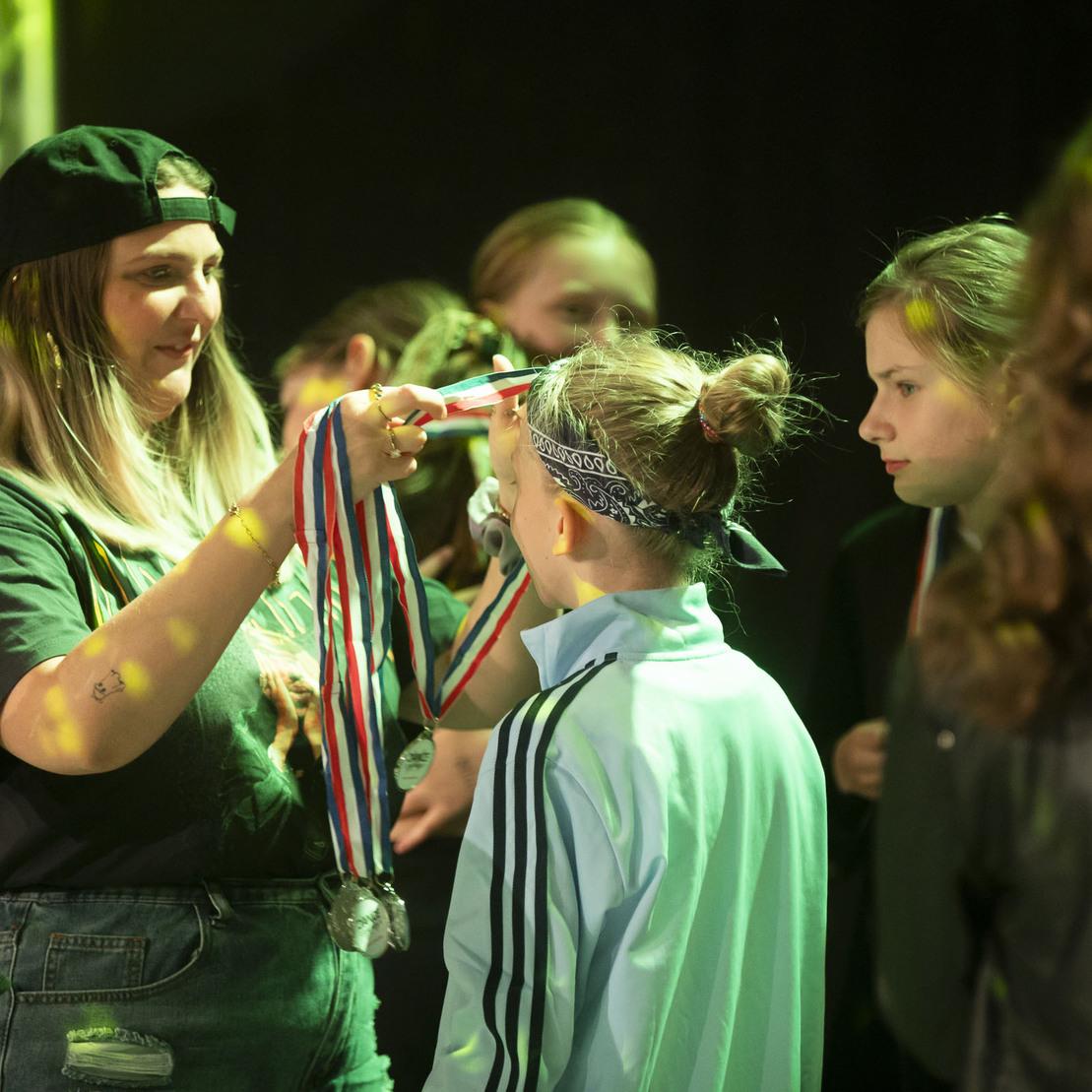 Eine Frau verleiht einer jungen Sportlerin eine Medaille, w&auml;hrend weitere Kinder aufmerksam zuschauen.