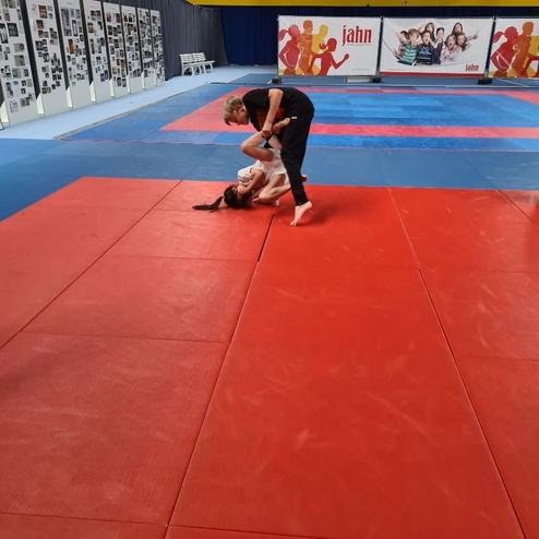 Junge in schwarzem Trainingstrikot &uuml;bt den Grappling-Move mit einem M&auml;dchen auf einer roten Matte in einem Sportraum.