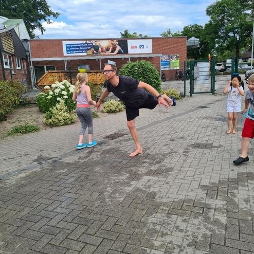 Ein Mann steht barfu&szlig; auf einem gepflasterten Platz und interagiert mit einem M&auml;dchen, w&auml;hrend Kinder im Hintergrund lachen.