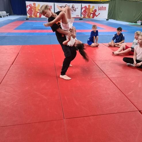 Zwei Kinder &uuml;ben eine Judotechnik auf einer roten Matte, w&auml;hrend andere Zuschauer aufmerksam zuschauen.