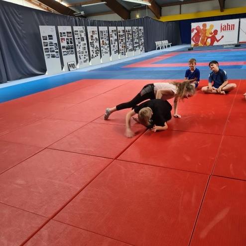 Kinder trainieren auf einer roten Mattenfl&auml;che; zwei Kinder f&uuml;hren eine &Uuml;bung aus, w&auml;hrend andere zuschauen.