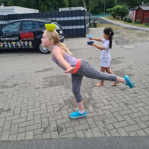 Ein M&auml;dchen balanciert eine Sch&uuml;ssel auf dem Kopf, w&auml;hrend ein anderes M&auml;dchen mit einer Sch&uuml;ssel in der Hand dasteht.