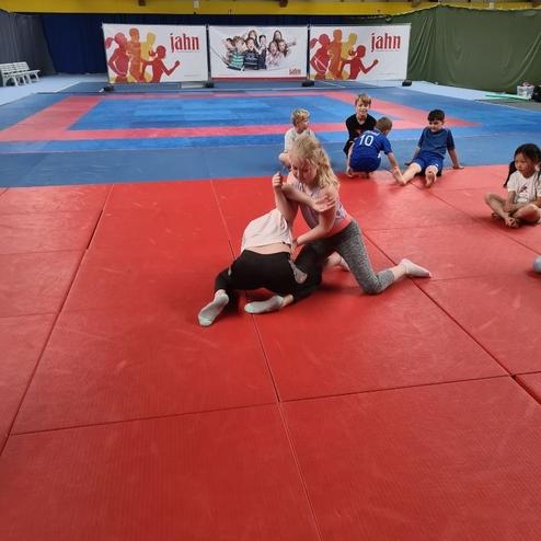 Zwei Kinder k&auml;mpfen auf einer roten Matte in einer Sporthalle, w&auml;hrend mehrere andere Kinder zuschauen.