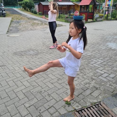 Zwei Kinder stehen auf einem Gehweg; eines balanciert eine Sch&uuml;ssel auf dem Kopf, das andere hebt ein Bein.