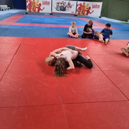 Zwei Kinder ringen auf einer roten Mattenfl&auml;che, w&auml;hrend andere Kinder im Hintergrund zuschauen.