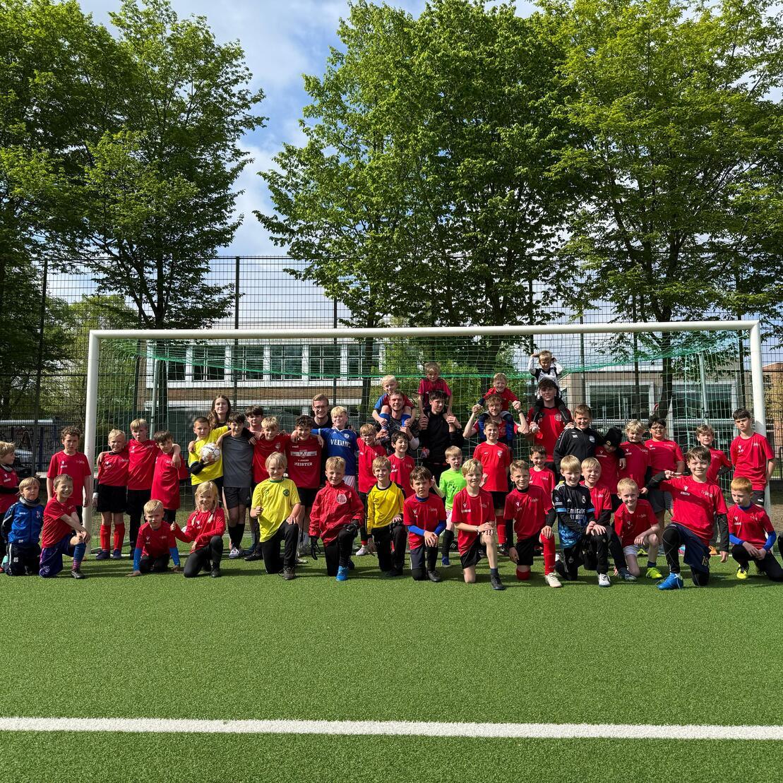 Gruppenfoto von Kindern in roten und gelben Trikots auf einem Fu&szlig;ballfeld vor dem Tor, umgeben von B&auml;umen.