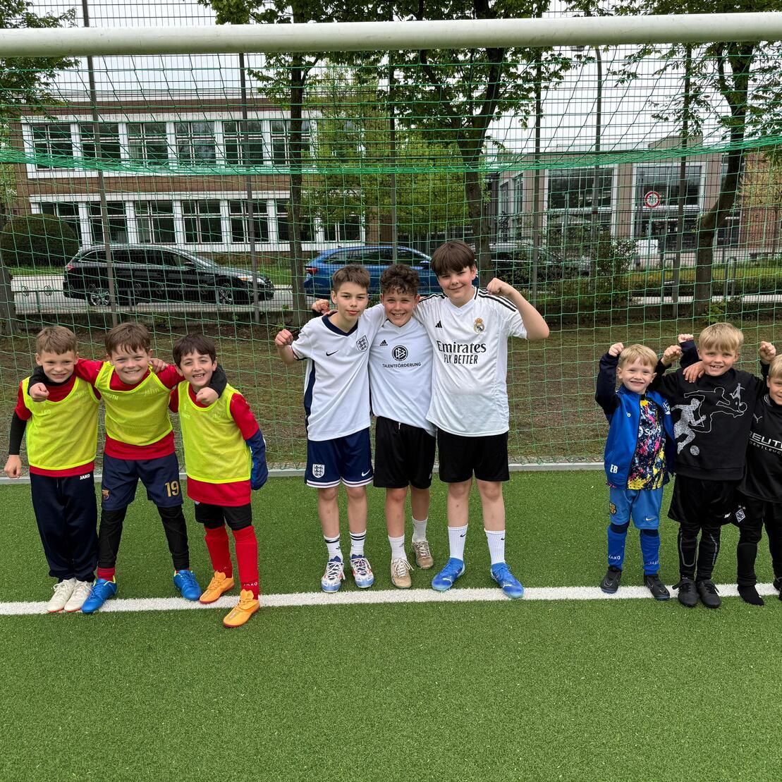Gruppe von sieben Kindern in Sportkleidung steht auf einem Fu&szlig;ballfeld vor einem Tor und l&auml;chelt in die Kamera.