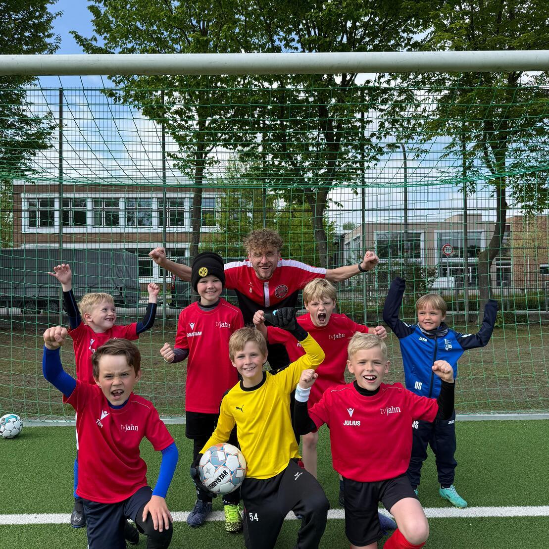 Gruppe von Kindern in Fu&szlig;balltrikots posiert fr&ouml;hlich vor einem Tor auf einem Sportplatz.