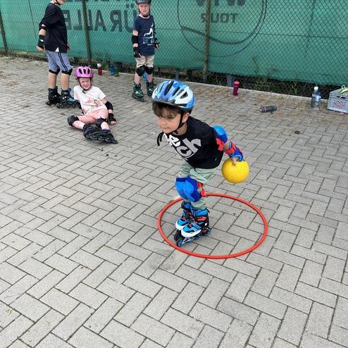 Kind mit Schutzhelm und Knieschonern rollt auf Inline-Skates, w&auml;hrend es einen Ball in einem roten Ring balanciert.