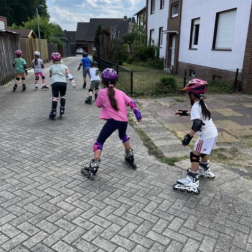 Sechs Kinder in Schutzkleidung skaten auf einer asphaltierten Stra&szlig;e neben Wohnh&auml;usern.
