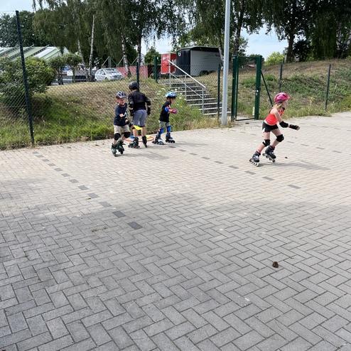 Vier Kinder in Schutzausr&uuml;stung fahren auf Inlineskates auf einem Pflasterplatz, umgeben von B&auml;umen und einem Zaun.