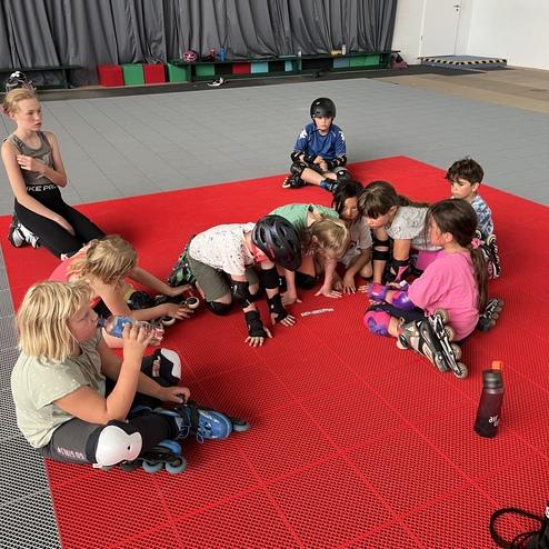 Gruppierung von Kindern in Schutzkleidung auf einer roten Fl&auml;che, konzentriert auf eine &Uuml;bung im Rollschuhlaufen.