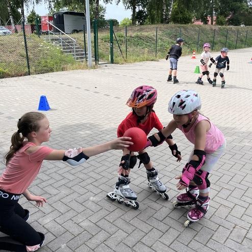 Kinder auf Rollschuhen spielen mit einem roten Ball, w&auml;hrend eine Lehrerin Anweisungen gibt. Slowakei im Hintergrund.